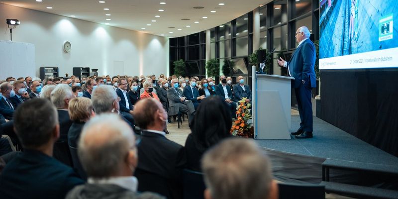 Publikum sitzt vor einer Bühne, grauhaariger Mann im Anzug steht auf der Bühne und hält ein Vortrag