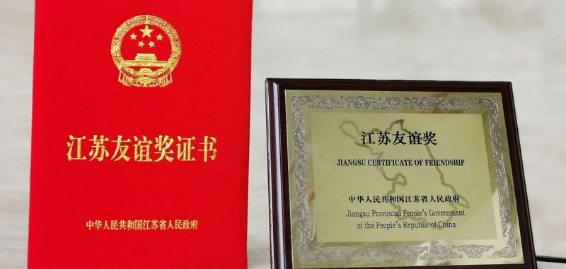 Freundschaftspreise, links rotes kleines Buch mit chinesischer goldener Aufschrift und rechts ein goldenes Bild mit holz Umrandung und schwarzer Schrift "Jiangsu Certificate of Friendship - Jiangsu Provincial People´s Government of the People´s Republic of China"